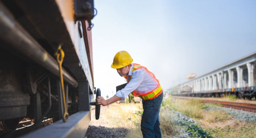 Trabalhador com capacete amarelo e colete refletivo ajusta componente em vagão de trem em área ferroviária.