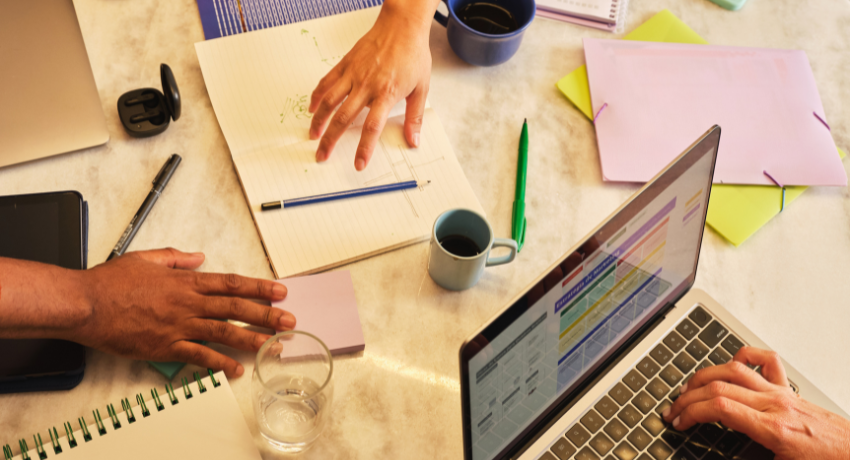 Mãos de profissionais trabalhando em mesa com laptop, anotações, café e materiais de planejamento.