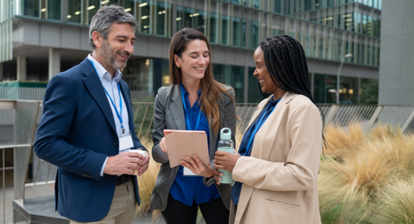 Três profissionais com crachás conversam ao ar livre diante de um escritório moderno; uma pessoa mostra um tablet e outra segura garrafa reutilizável.