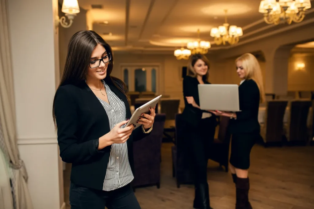 Equipe de eventos em um salão elegante: mulher com tablet na mão lê informações de um evento enquanto outras pessoas analisam documentos, cenário que sugere estratégia para transformar visitantes em solicitações de orçamento. Sala com iluminação e mesas ao fundo.